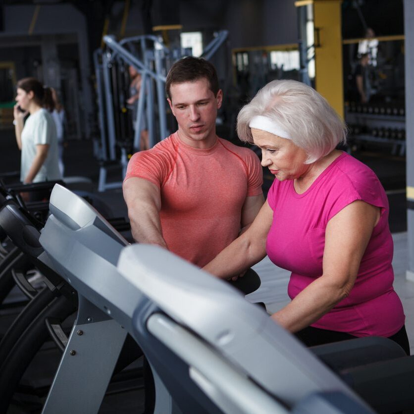 A trainer assists an elderly woman on a treadmill.